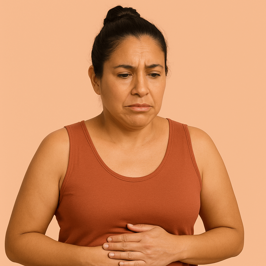 Mexican woman in her 40s holding her abdomen and looking slightly uncomfortable against a peach background, illustrating abdominal cramps during menopause for an article on Abdominal Cramps and Menopause: What’s Going On and How to Find Relief.